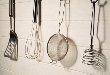 Spatula, whisk, masher and strainer hanging against a rustic white wall in restaurant or home kitchen. Kitchen utensils made of stainless steel or metal. Set of tools for cooking food. Kitchenware on