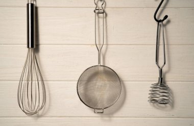 Whisk, masher and strainer hanging against a rustic white wall in restaurant or home kitchen. Kitchen utensils made of stainless steel or metal. Set of tools for cooking food. Kitchenware on drainer