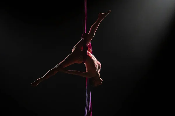 Female circus gymnast hanging upside down on aerial silk and demonstrates stretching. Young woman performs tricks at height on a red silk fabric. Difficult acrobatic stunts on black background with