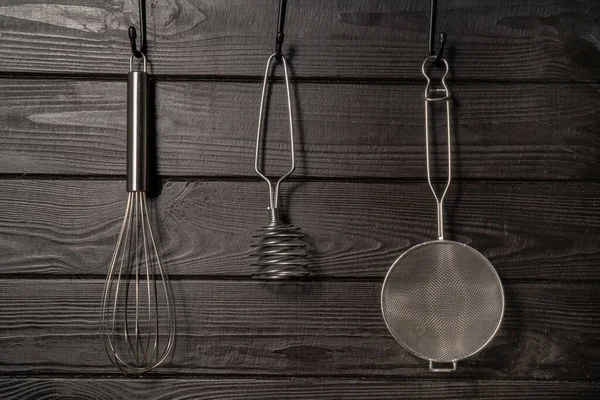 Whisk, masher and strainer hanging against a rustic grey wall in restaurant or home kitchen. Kitchen utensils made of stainless steel or metal. Set of tools for cooking food. Kitchenware on drainer