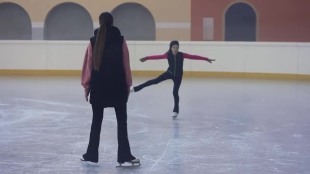 スケート場のアイスアリーナで小さな女の子が1本足で滑り 回転します 女子フィギュアスケートコーチは生徒を賞賛し 彼女を賞賛します 軽競技場でフィギュアスケートの練習 — ストック動画