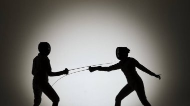 Siyah siluetlerin yan görüntüsü çapraz saplı iki genç kadın kılıç ustası. Beyaz üniformalı ve koruyucu miğferli sporcular arka plandaki karanlık stüdyoda düello yapıyorlar. Kucağında çember tutan kızlar