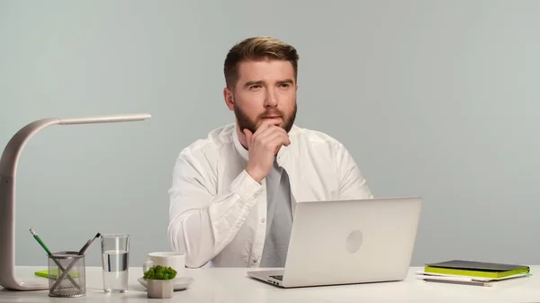 Young bearded man in white shirt thinking intently. Worker or student sitting at white table with laptop at home or in office. Freelancer works remotely. Remote education. Online work during