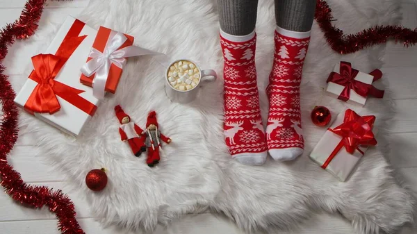 Female legs in red warm socks with white deer and snowflakes on white fur. Tinsel, gift boxes with bows, Christmas balls and toys are laid out on floor. Cup of hot cocoa with marshmallows. Winter