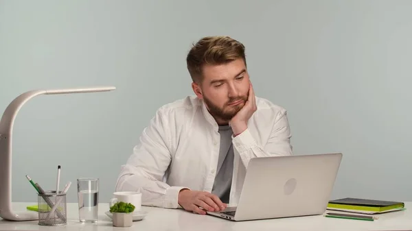 白いシャツを着た若い髭の男がノートパソコンの画面を見て考えている 労働者や学生は 自宅やオフィスでノートパソコンと白いテーブルに座っている フリーランスはリモートで働いています 遠隔教育 オンライン — ストック写真