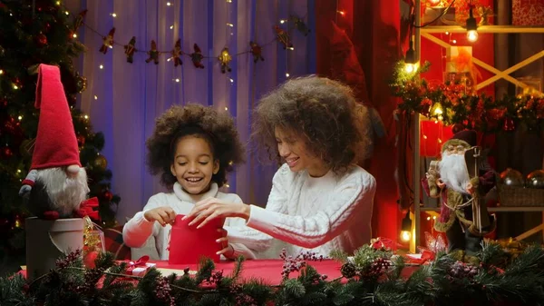 African American mom and little daughter packing letter for Santa Claus in red envelope. Woman and girl sitting near Christmas tree against home room decorated for festive night. Happy family portrait