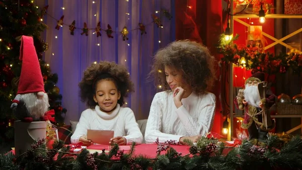African American mom and little daughter reading a letter written for Santa Claus. Woman and girl sitting near Christmas tree against home room decorated for festive night. Happy family portrait. New