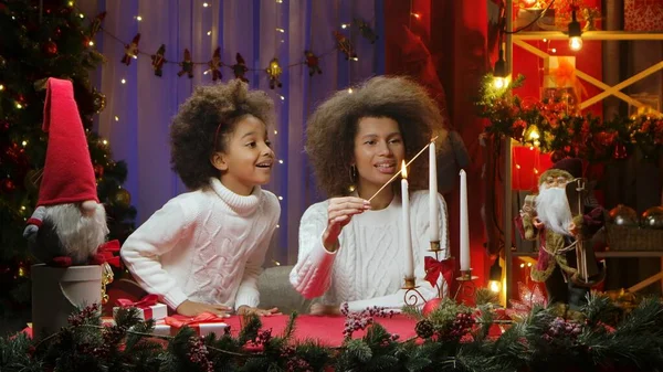 African American mom and little daughter burning white candles with wow delight emotions. Woman and girl sitting near Christmas tree against home room decorated for festive night. Happy family