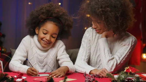 African American mom and little daughter writing letter to Santa Claus and smiling. Woman and girl in white sweaters sitting against home room. Happy family portrait. New Year and Christmas