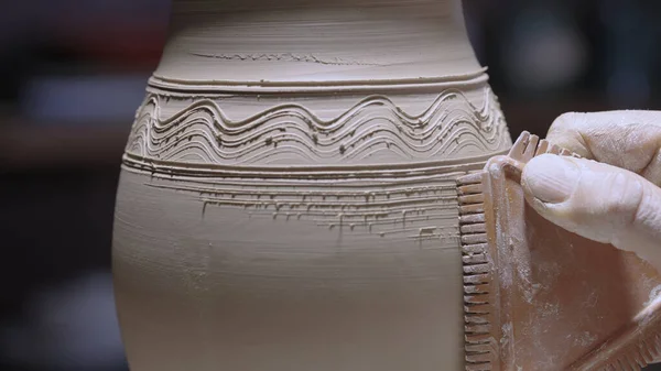 Male hands of a potter carve designs on a soft white clay pot with pottery tools. A man decorates a pot with a serrated scraper, drawing lines and waves. Unfired earthenware in a workshop close up