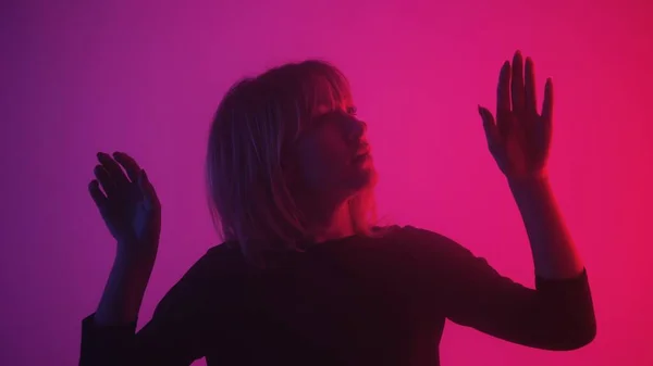 Young woman touches the glass with her palms. Blonde examines glass and slides her hands over it with interest and surprise. Model poses in dark studio with pink purple lights and smoke. Close up