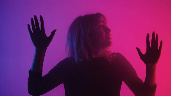 Frightened young woman touches glass with palms. Blonde examines glass and slides her hands over it with upset and afraid. Model poses in dark studio with pink purple lights and smoke. Close up