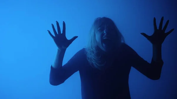 Young woman touches glass with palms and and screams in fear and horror. Woman face behind glass conveys emotions afraid, fear, fright, dread. Model poses in dark studio with white blue lights and