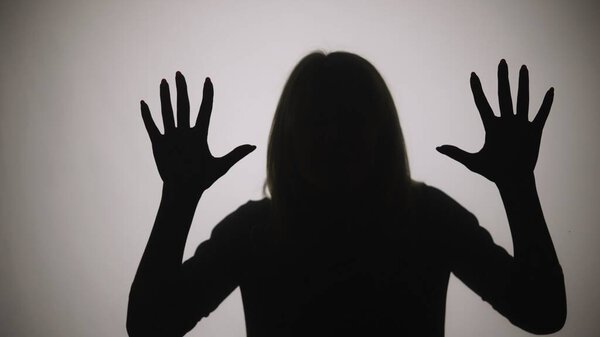 Black silhouette of a woman touching the glass with her palms and looking at the camera. The outline of a woman against the background of a window with white matte light. Model poses in studio with