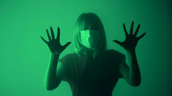 戴着医疗面罩的女人看着相机 在玻璃上摸她的手掌 一个心烦意乱的女人用手在黑暗的工作室里 在绿色柔和的灯光和浓烟的背景下触摸着玻璃 — 图库照片