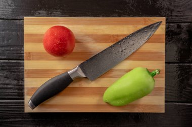 Yeşil tatlı biber, kırmızı domates ve tahta kesme tahtasında keskin metal bıçak. Taze çiğ sebzeler ve koyu gri ahşap bir masada yağmur damlalarından ıslanmış bir bıçak. Sağlıklı bir salata için mevsimlik sebzeler