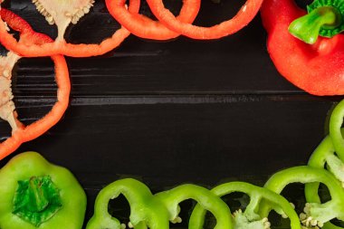 Yeşil ve kırmızı biber, siyah ahşap bir masa ya da çalışma alanı yazılı mutfak masası yüzeyinde halka şeklinde kesilmiş. Yuvarlak çerçeve. Dilimlenmiş çekirdekli dolgun dolma biber parçalarının üst görüntüsü.