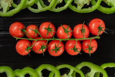 Bir demet olgun kırmızı kiraz domatesleri ve siyah tahta üzerinde doğranmış yeşil tatlı biber. Domateslerin üst tarafı ve su damlalarından ıslanan dilimlenmiş dolma biber parçaları. Taze sebzeler