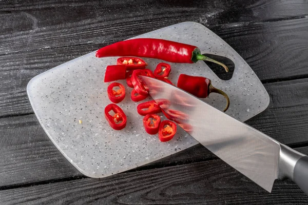 Sharp metal knife slicing rings of red hot pepper lying on a gray plastic kitchen board. Spicy chili pepper chopped slices on a dark gray wooden table. Cooking in a restaurant. Condiment, spicy spice