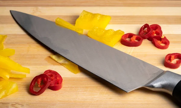 Sliced pieces of yellow sweet pepper, red hot chili pepper and a metal sharp knife on a wooden board. Juicy pulp of yellow bell pepper and red cayenne pepper on cutting board. Background of ripe