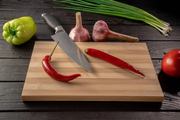 Kırmızı kırmızı kırmızı kırmızı biberler ve tahta kesme tahtasının üzerinde keskin metal bir bıçak. Tatlı biber, yeşil soğan sapı, sarımsak ve domates. Koyu gri ahşap bir masada yatıyor. Taze çiğ sebze ve baharat.