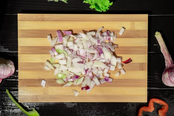 Top view of chopped purple onion on a wooden kitchen board. Garlic, lettuce green, red and green sweet pepper slices on a wet black wooden table surface. Close up of vegetables sliced for salad