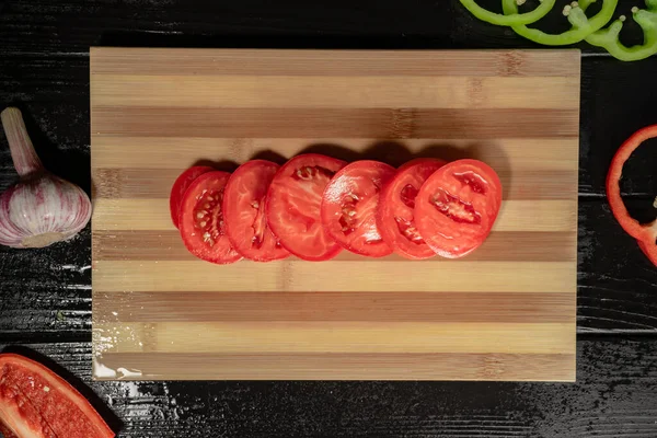 Top view of red ripe tomato is cutting into rings on a wooden kitchen board with water drops. Close up of sliced tomato, garlic, round slices green and red sweet pepper on black surface of wooden wet