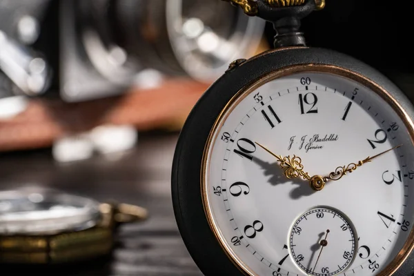 Silver antique swiss pocket watch with gold hands and vintage film camera in background. An old round dial of a pocket watch on a dark wooden table and blurry camera lens with aperture. Time flow