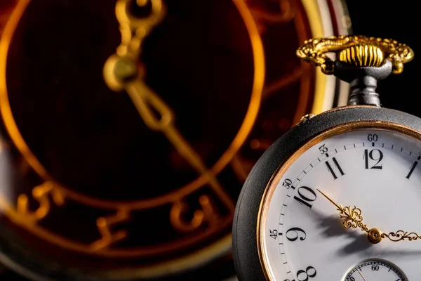 Silver antique pocket watch with gold hands and vintage table clock in blurry background. An old round white dial of pocket watch and brown face of table clock with hands on dark background. Close up