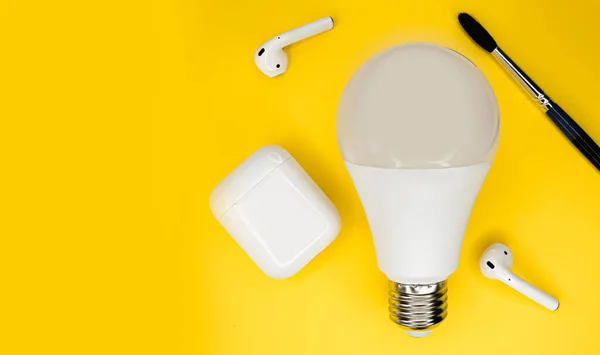 Una hermosa bombilla blanca sobre un fondo minimalista. Idea de lámpara. Auriculares inalámbricos y pincel. — Foto de Stock