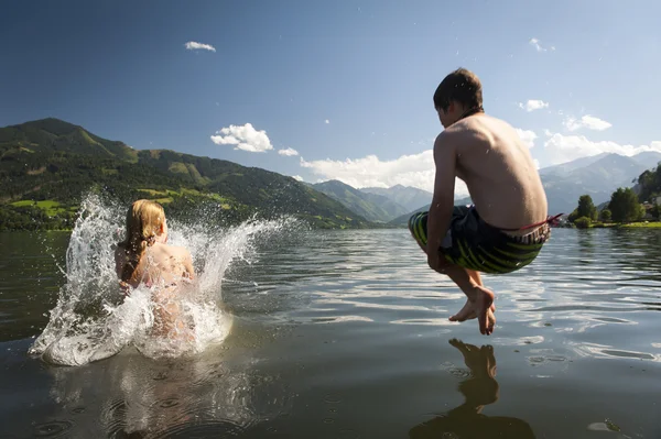 Girl already in the splashing water and boy in the air while they where jumoing into a lake, with nice nature and mountains in the back — Stock Photo, Image