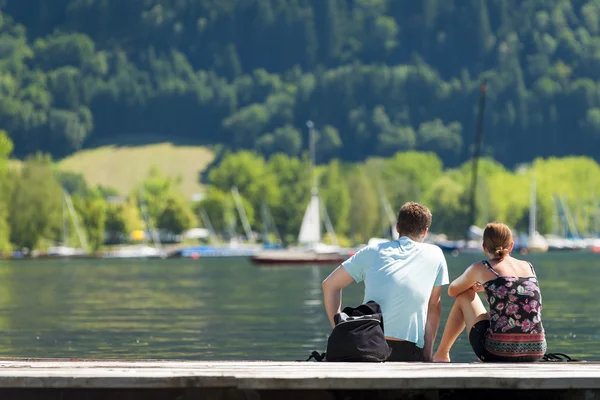 Vista trasera de una joven pareja sentada a orillas del lago bajo el sol del verano admirando los barcos amarrados y los yates — Foto de Stock