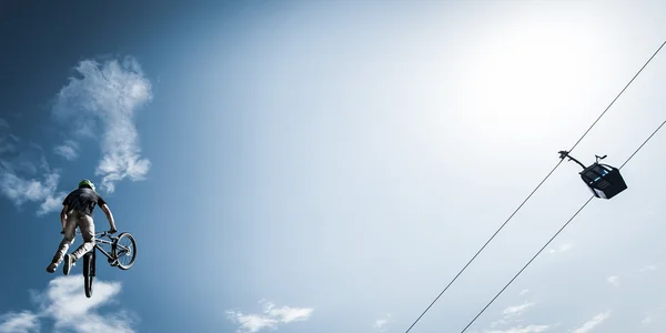 Joven motociclista de acrobacias salta junto al teleférico con el cielo azul nublado en la parte posterior —  Fotos de Stock