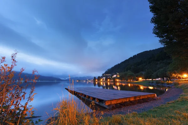 Sunset over a tranquil lake with a wooden jetty and colourful lights lining the shore — Stock Photo, Image