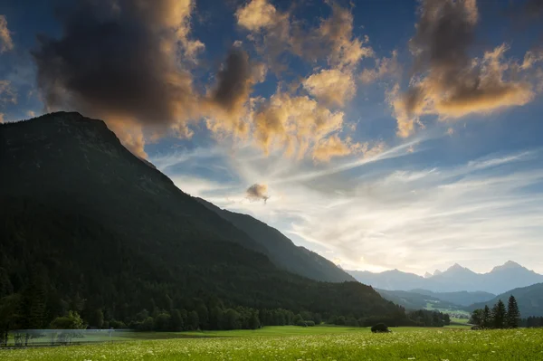 Exuberante prado verde en un valle de montaña al atardecer rodeado de picos silouetted bajo nubes naranjas brillantes — Foto de Stock