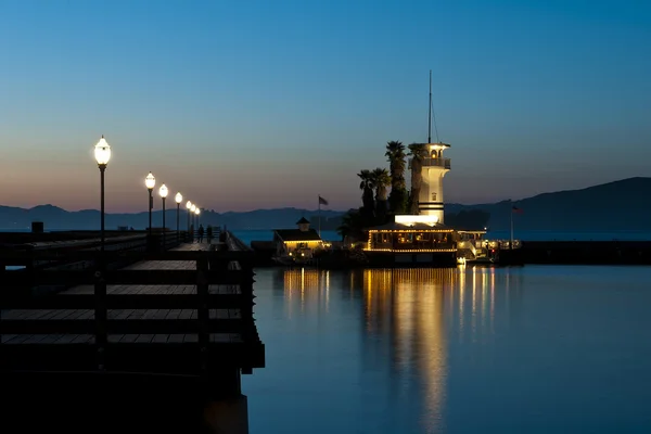 Faro de torre iluminada con restaurante junto a una pasarela al amanecer con un bonito espejo en el agua — Foto de Stock