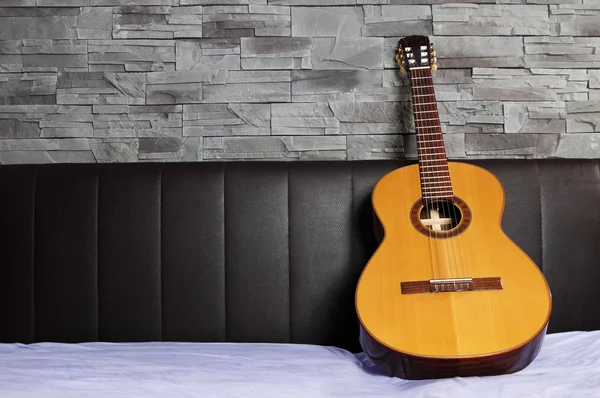 Classical guitar lying on the bed in front of a brown leather back and a stone wall — Stock Photo, Image