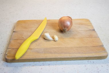 Derisindeki sarı soğan, soyulmamış bir diş sarımsak ve tahtanın üzerinde duran sarı bir bıçak..