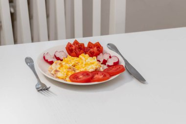 Sağlıklı yiyecekler. Beyaz bir tabakta çırpılmış yumurta ve salam. Etrafta sebze var. Turp ve domates..