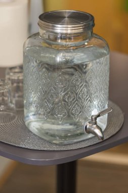 Ev yapımı cam su kabı masanın üzerinde duruyor. Konteynırda su musluğu var..