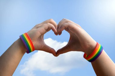 Igbt, eşcinsel aşk ve homoseksüel ilişkiler konsepti - gay gururlu erkek ellerine yakın gökkuşağı farkındalığı bileklikleri mavi arka planda kalp hareketi gösteriyor.