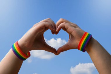 Igbt, eşcinsel aşk ve homoseksüel ilişkiler konsepti - gay gururlu erkek ellerine yakın gökkuşağı farkındalığı bileklikleri mavi arka planda kalp hareketi gösteriyor.