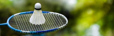 Beyaz kremalı badminton topu ve açık havada bulanık ağaçlar ve halka açık park geçmişi olan mavi badminton raketi, açık alanda eğlence amaçlı spor aktiviteleri konsepti..