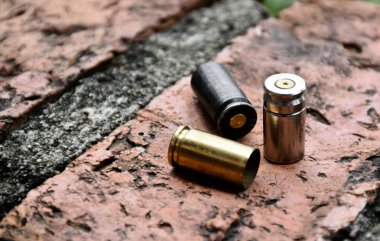 Tuğla zeminde 9mm 'lik tabanca mermileri ve mermi kovanları, yumuşak ve seçici bir şekilde siyah mermi kovanına odaklanma, olay yerindeki bir cinayet davasında kilit bir kanıtı arama kavramı.