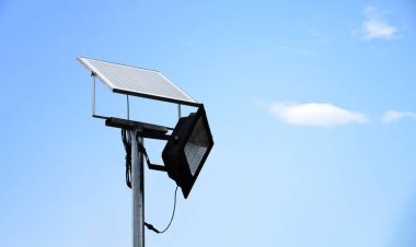 Mini photovoltaic or solar cell panel and floodlight led installed on metal pole against blue sky to store and save the energy from the sun to use the light at night around the house or the park.