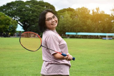 Portrain of asian middle aged woman holding badminton racket, standing and smiling in the green field to play outdoor badminton, concept for to be strong and healthy woman by exercising afterwork.
