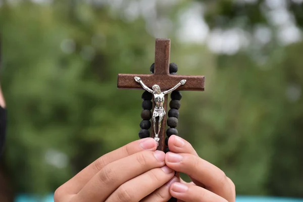 Wooden cross and wooden rosary are held in hands of young asian Catholic prayer while praying in the temple park area.