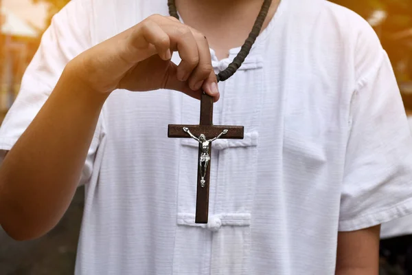 Wooden cross and wooden rosary are held in hands of young asian Catholic prayer while praying in the temple park area.