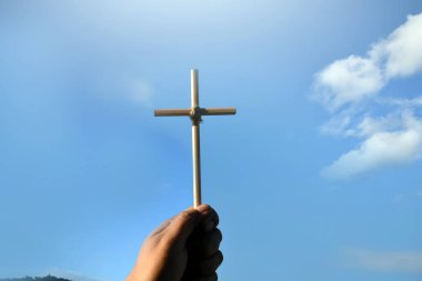 Bulutlu ve mavi gökyüzü arkaplanıyla el ele tutuşan küçük ahşap haç, aşk, umut, doğruluk, inanç, İsa 'ya inan, yumuşak ve seçici odaklanma konsepti.