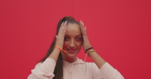 Chica joven y moderna con auriculares sonriendo posando. Bastante hembra en auriculares sobre fondo rojo con espacio para copiar — Vídeos de Stock